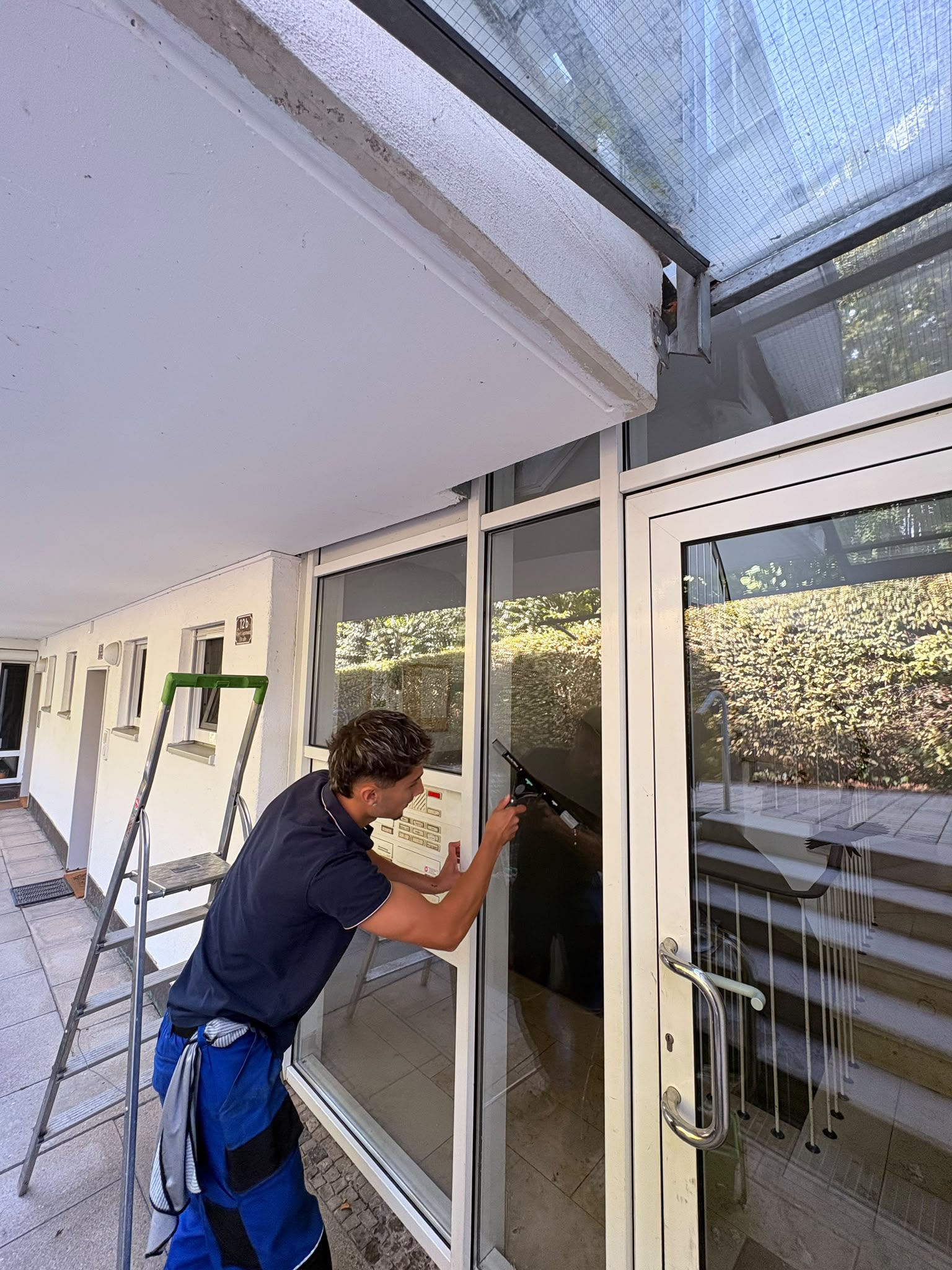 Mitarbeiter reinigt die Glasflaechen eines Hauseingangs, daneben eine Leiter
